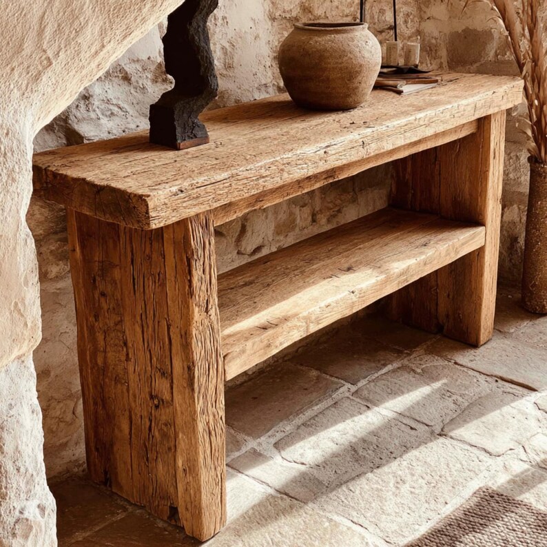 Op de afbeelding: Een rustieke houten console met een onderste plank. Het hout toont natuurlijke nerven en textuur. Een donkere sculptuur en een keramische vaas staan op het tafelblad. De console staat tegen een stenen muur. Ongeveer 150 cm lang.