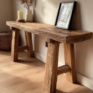 May include: A weathered wooden bench with a natural finish. The bench features a thick, rectangular top supported by angled legs. A framed picture, a candle, and a vase with dried plants are on the bench. The bench is approximately 150 cm long.