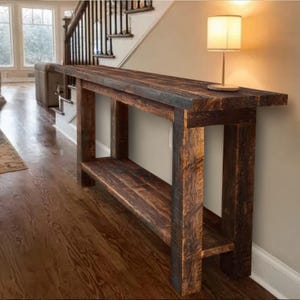 Op de afbeelding: Een rustieke, donker gebeitste houten console met een onderste plank. De tafel heeft een rechthoekig blad en stevige poten, die de natuurlijke nerf van het hout laten zien. Een kleine lamp staat op de tafel. De tafel staat tegen een neutrale muur.
