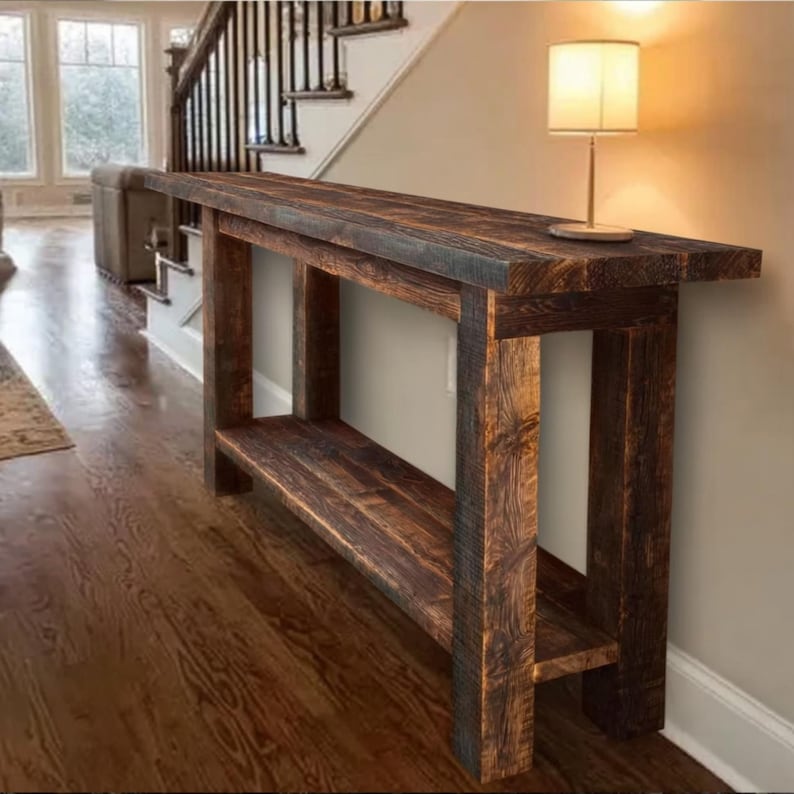 Op de afbeelding: Een rustieke, donker gebeitste houten console met een onderste plank. De tafel heeft een rechthoekig blad en stevige poten, die de natuurlijke nerf van het hout laten zien. Een kleine lamp staat op de tafel. De tafel staat tegen een neutrale muur.