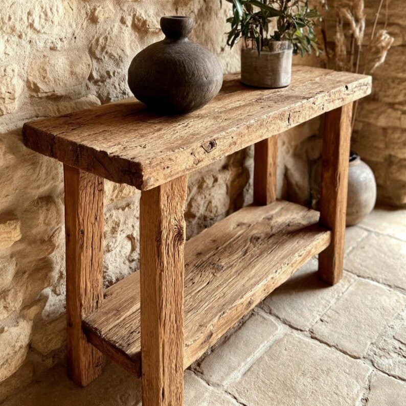 Op de afbeelding: Een rustieke houten console met een onderste plank. De tafel is gemaakt van gerecycled hout, met natuurlijke knopen en nerven. Een donkere, ronde vaas en een kleine plant in pot staan op het tafelblad. De tafel is ongeveer 122 cm lang.