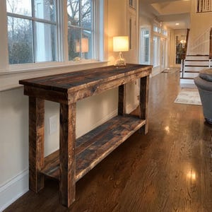 Op de afbeelding: Een rustieke houten console met een donkerbruine afwerking. De tafel heeft een onderste plank en is geconstrueerd met zichtbare houtnerf en spijkerkoppen. Op het oppervlak van de tafel staat een kleine lamp. De tafel is ongeveer 152 cm lang.