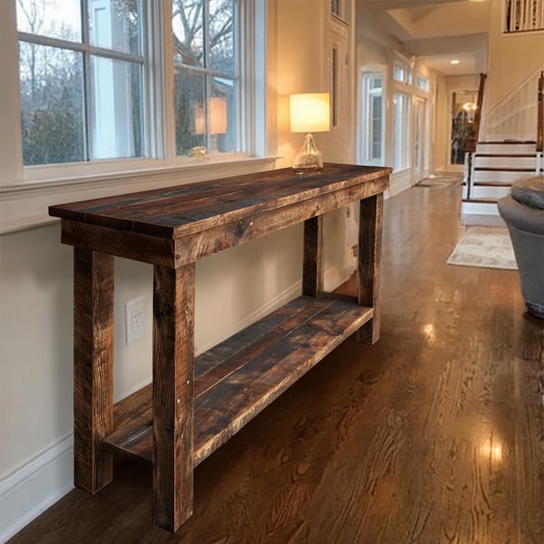 Op de afbeelding: Een rustieke houten console met een donkerbruine afwerking. De tafel heeft een onderste plank en is geconstrueerd met zichtbare houtnerf en spijkerkoppen. Op het oppervlak van de tafel staat een kleine lamp. De tafel is ongeveer 152 cm lang.