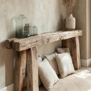 Puede incluir: Una consola de madera rústica con un acabado envejecido. La mesa presenta una gruesa tapa rectangular sostenida por patas cuadradas y robustas. Frascos de vidrio decorativos y un jarrón con plantas secas se encuentran sobre la mesa, con varias almohadas de color neutro debajo.