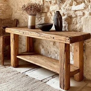 Op de afbeelding: Een rustieke houten console met een onderste plank. De tafel is gemaakt van gerecycled hout, met natuurlijke nerven en knopen. Een vaas met gedroogde bloemen, een zwarte kom en een donkere vaas staan erop. Het ontwerp van de tafel is eenvoudig en stevig.
