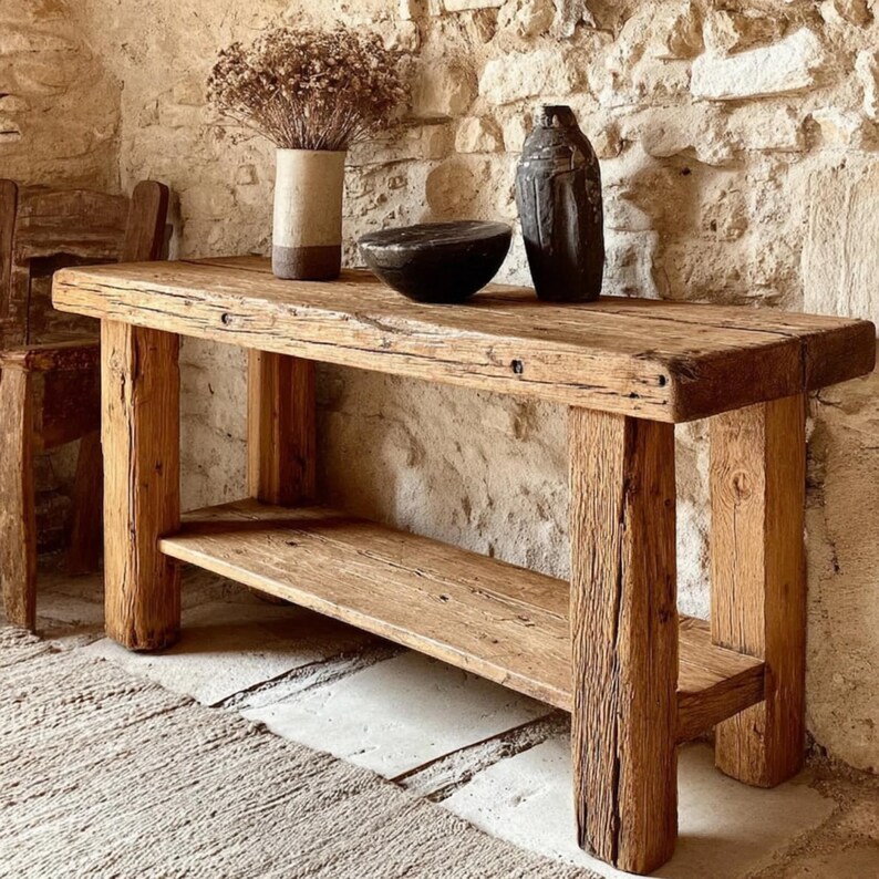 Op de afbeelding: Een rustieke houten console met een onderste plank. De tafel is gemaakt van gerecycled hout, met natuurlijke nerven en knopen. Een vaas met gedroogde bloemen, een zwarte kom en een donkere vaas staan erop. Het ontwerp van de tafel is eenvoudig en stevig.