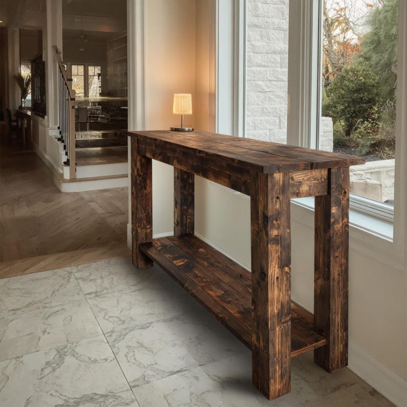 Op de afbeelding: Een rustieke, donkerbruine houten console met een onderste plank. De tafel is gemaakt van dik, gerecycled hout, dat de natuurlijke nerf en knopen laat zien. Hij staat voor een groot raam, met een kleine lamp erop.