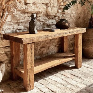 Op de afbeelding: Een rustieke houten console met een onderste plank. De tafel is gemaakt van gerecycled hout en toont natuurlijke nerven en textuur. Een klein beeld en een donkere vaas staan erop. De tafel staat tegen een stenen muur.