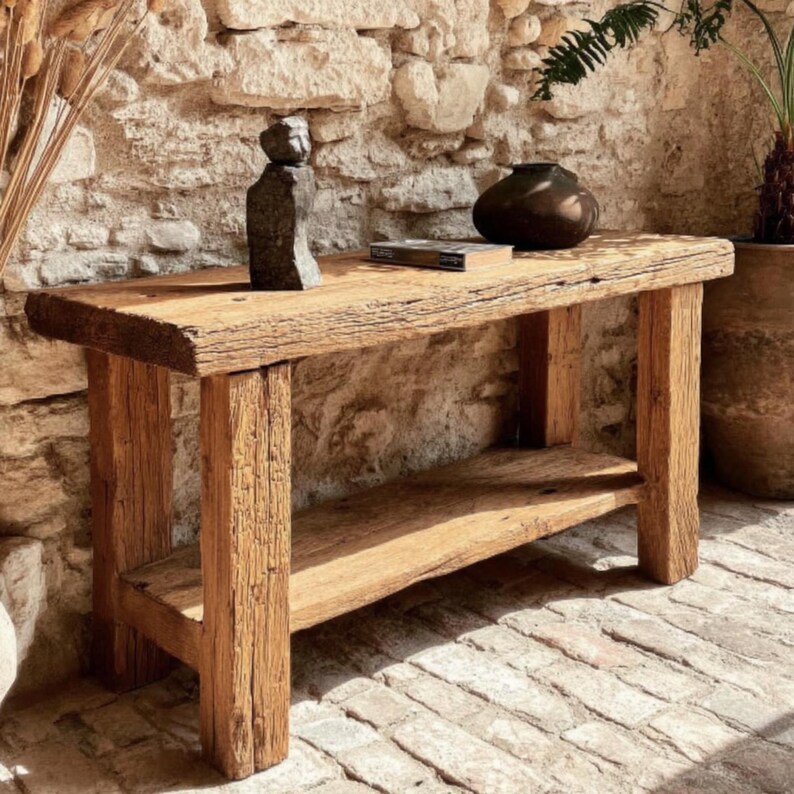 Op de afbeelding: Een rustieke houten console met een onderste plank. De tafel is gemaakt van gerecycled hout en toont natuurlijke nerven en textuur. Een klein beeld en een donkere vaas staan erop. De tafel staat tegen een stenen muur.