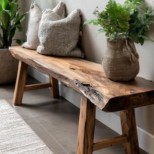 May include: A wooden bench with a natural, live edge. The bench has a light brown finish and wooden legs. Two textured grey cushions are on the bench, and a woven basket with greenery sits on the right. A potted plant is visible on the left.