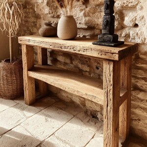Op de afbeelding: Een rustieke houten console met een onderste plank. De tafel is gemaakt van gerecycled hout en heeft een natuurlijke, verweerde uitstraling. Vazen en een sculptuur staan op het tafelblad. De tafel staat tegen een stenen muur.