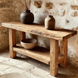 Op de afbeelding: Een rustieke houten console met een onderste plank. Twee donkere vazen en een geweven kom staan op de tafel. De tafel is gemaakt van gerecycled hout en heeft een natuurlijke afwerking. De achtergrond is een stenen muur.