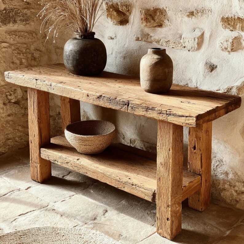 Op de afbeelding: Een rustieke houten console met een onderste plank. Twee donkere vazen en een geweven kom staan op de tafel. De tafel is gemaakt van gerecycled hout en heeft een natuurlijke afwerking. De achtergrond is een stenen muur.
