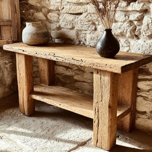 Op de afbeelding: Een rustieke houten console met een onderste plank en dikke poten. De tafel is gemaakt van lichtbruin hout en heeft een natuurlijke, verweerde uitstraling. Twee keramische vazen en gedroogde planten worden op de tafel tentoongesteld. De achtergrond is een stenen muur.