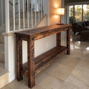 Op de afbeelding: Een rustieke houten console met een onderste plank. De tafel is gemaakt van donker gebeitst hout, met de natuurlijke nerf en knopen. De tafel is ongeveer 183 cm lang, 41 cm breed en 76 cm hoog.