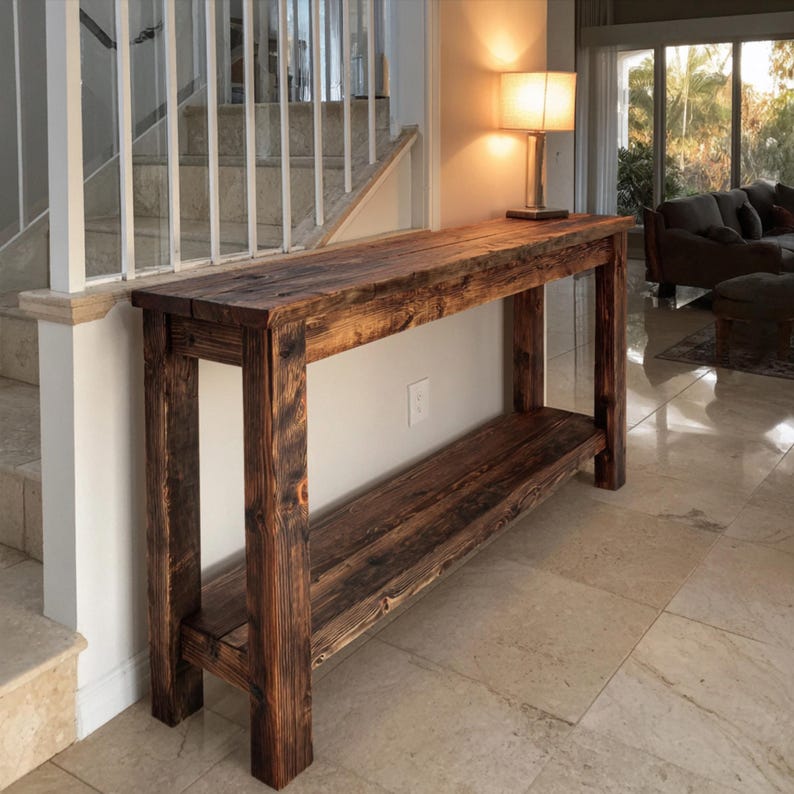 Op de afbeelding: Een rustieke houten console met een onderste plank. De tafel is gemaakt van donker gebeitst hout, met de natuurlijke nerf en knopen. De tafel is ongeveer 183 cm lang, 41 cm breed en 76 cm hoog.