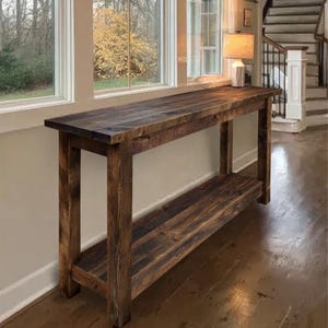 Op de afbeelding: Een rustieke, donker gebeitste houten consoletafel met een onderste plank. De tafel heeft een rechthoekige bovenkant en vier stevige poten. De houtnerf is zichtbaar, wat bijdraagt aan de natuurlijke esthetiek. De tafel is ongeveer 183 cm lang.