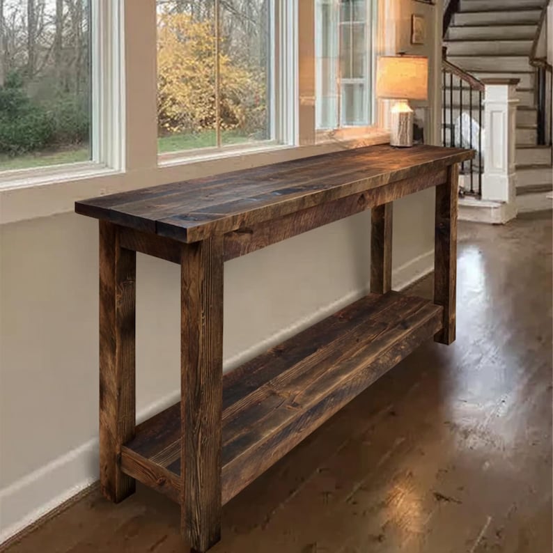 Op de afbeelding: Een rustieke, donker gebeitste houten consoletafel met een onderste plank. De tafel heeft een rechthoekige bovenkant en vier stevige poten. De houtnerf is zichtbaar, wat bijdraagt aan de natuurlijke esthetiek. De tafel is ongeveer 183 cm lang.