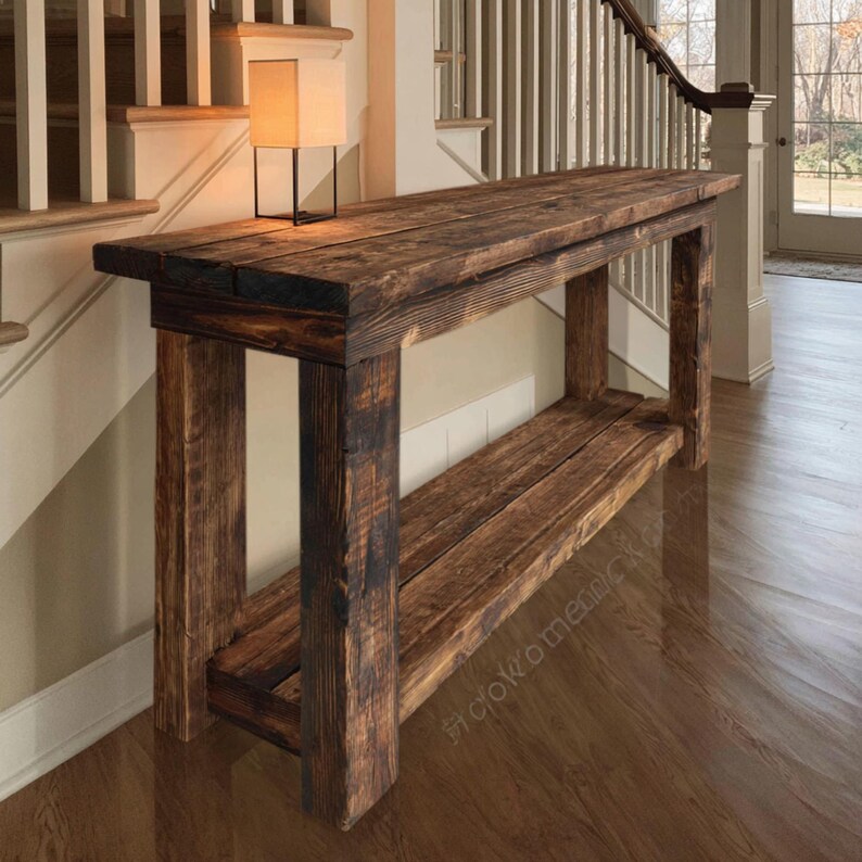 Op de afbeelding: Een rustieke houten console met een onderste plank. De tafel is gemaakt van donker gebeitst hout, dat de natuurlijke nerfstructuur laat zien. Het ontwerp heeft een rechthoekig blad en stevige poten, perfect voor een entree of woonkamer. Een kleine lamp staat op de tafel.
