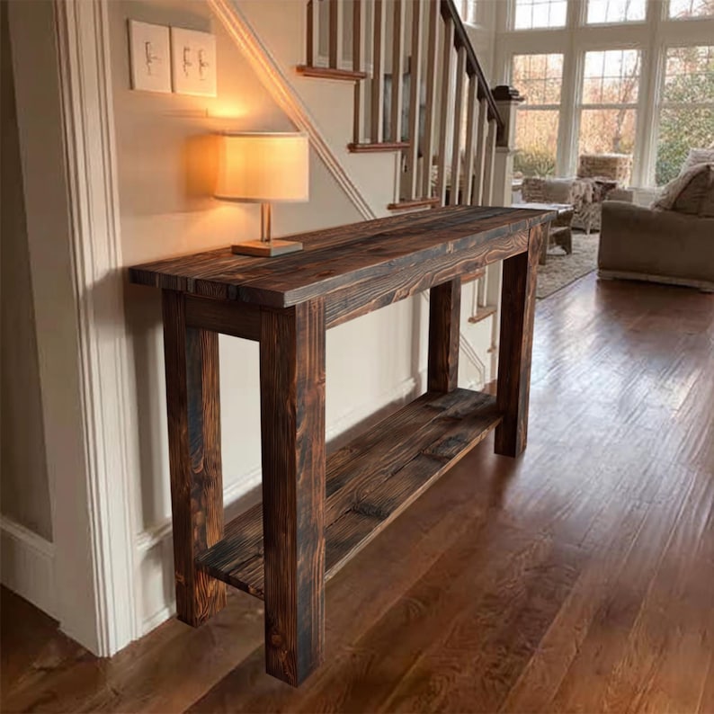Op de afbeelding: Een rustieke, donker gebeitste houten consoletafel met een onderste plank. De tafel heeft een rechthoekig blad en stevige poten. Op de tafel staat een kleine lamp met een beige kap. De tafel staat in een lichte kamer met hardhouten vloeren.