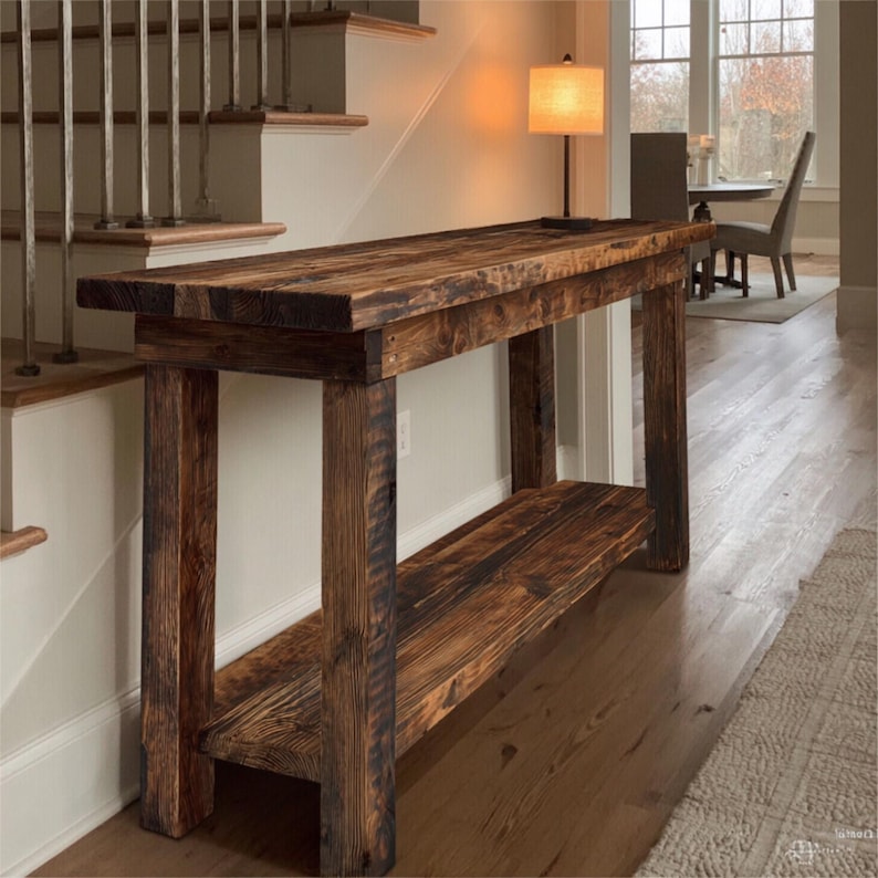 Op de afbeelding: Een rustieke, donker gebeitste houten console tafel. De tafel heeft een lange, rechthoekige bovenkant met een onderste plank en stevige, vierkante poten. De houtnerf is zichtbaar, wat bijdraagt aan de natuurlijke, handgemaakte uitstraling. De tafel is ongeveer 152 cm lang.