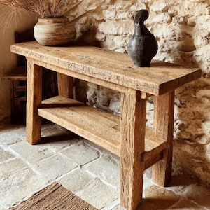 Op de afbeelding: Een rustieke houten console met een onderste plank. De tafel is gemaakt van lichtbruin, verweerd hout en heeft een natuurlijke, onafgewerkte uitstraling. Een donkere vaas staat erop en een decoratieve vaas met gedroogd gras is ook aanwezig.