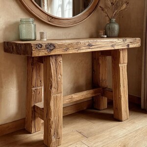 May include: A reclaimed wood console table with a natural finish. The table has a thick, rectangular top supported by square legs. A round mirror and decorative items sit on the table. The table's design creates a warm, inviting aesthetic. The table is made of reclaimed wood.