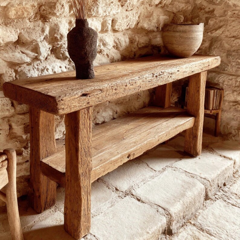 Op de afbeelding: Een rustieke houten console met een onderste plank. De tafel is gemaakt van gerecycled hout, met natuurlijke nerven en knopen. Een donkerbruine vaas met gedroogde stelen en een lichtbruine kom staan op de tafel. De tafel is 152 cm lang.