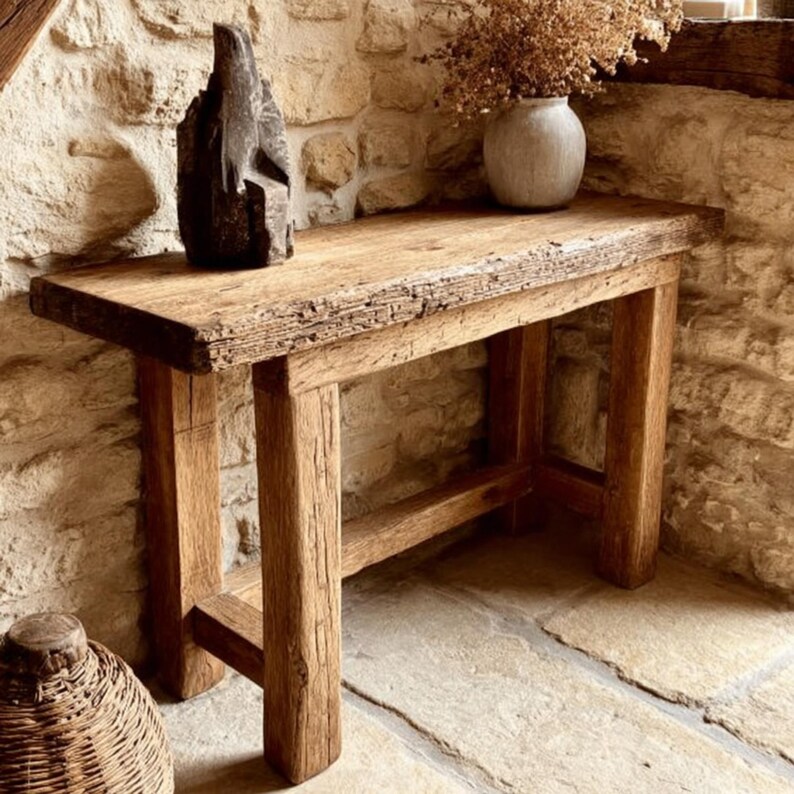 Op de afbeelding: Een rustieke houten console met een natuurlijke afwerking. De tafel heeft een dik, verweerd blad en stevige poten. Een donkere sculptuur en een vaas met gedroogde bloemen staan op de tafel. De tafel staat tegen een stenen muur.