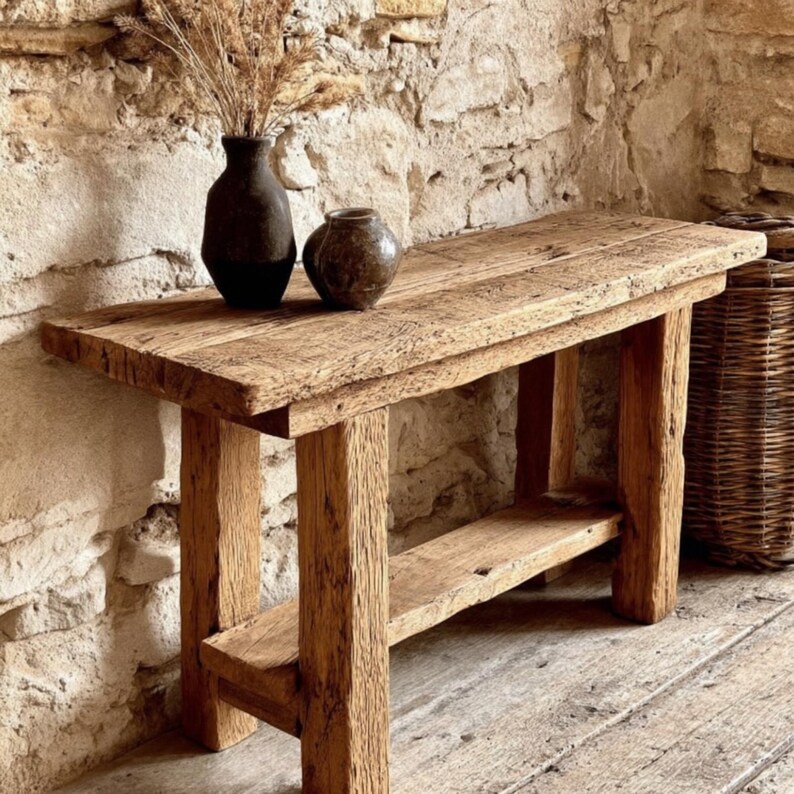 Op de afbeelding: Een rustieke houten console met een onderste plank. Twee donkerbruine keramische vazen en gedroogde grassen staan op het tafelblad. De tafel staat tegen een stenen muur en een houten vloer. De tafel is ongeveer 120 cm lang.