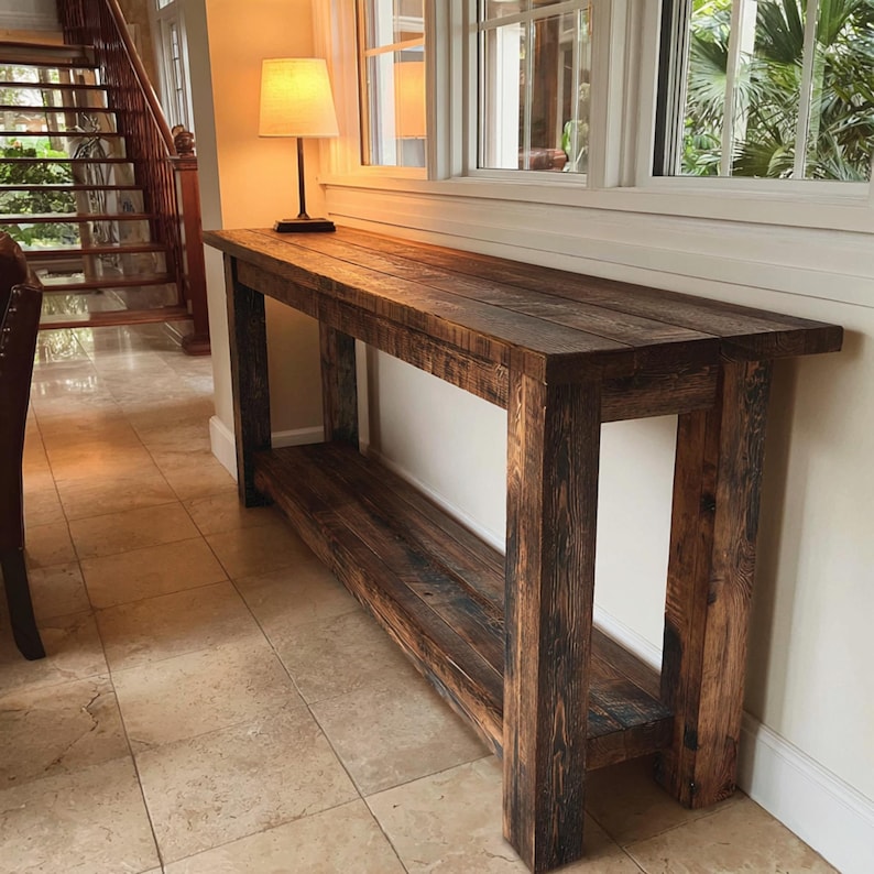 Op de afbeelding: Een rechthoekige houten console met een onderste plank, in donkerbruine kleur. De tafel is gemaakt van gerecycled hout met zichtbare nerven en knopen. Hij staat tegen een witte muur, met een lamp erop.