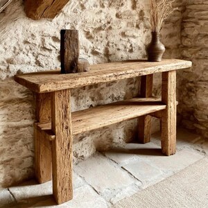 Op de afbeelding: Een rustieke houten console met een onderste plank, gemaakt van gerecycled hout. De tafel heeft een natuurlijke, verweerde afwerking en is versierd met een kleine vaas met gedroogde grassen en een houten sculptuur. De tafel is ongeveer 152 cm lang.