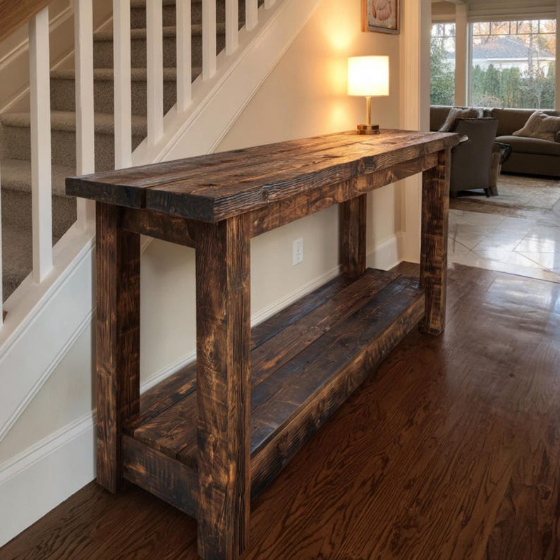 Op de afbeelding: Een rustieke, donker gebeitste houten consoletafel. De tafel heeft een lange, rechthoekige bovenkant en een onderste plank, ondersteund door dikke, vierkante poten. De houtnerf is zichtbaar, wat bijdraagt aan het natuurlijke, verouderde uiterlijk van de tafel. De tafel is ongeveer 152 cm lang.