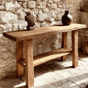 Op de afbeelding: Een rustieke houten console met een onderste plank. De tafel is gemaakt van gerecycled hout, met natuurlijke nerven en knopen. Een donkerbruine vaas met gedroogde planten en een houten sculptuur sieren het tafelblad. De tafel is ongeveer 150 cm lang en heeft dikke, vierkante poten.