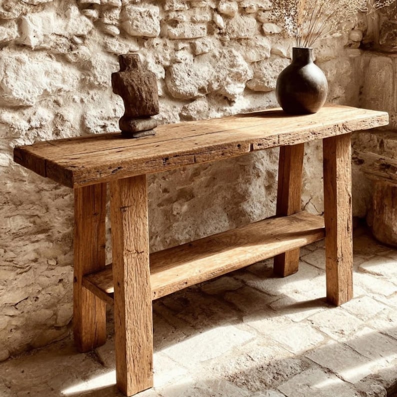 Op de afbeelding: Een rustieke houten console met een onderste plank. De tafel is gemaakt van gerecycled hout, met natuurlijke nerven en knopen. Een donkerbruine vaas met gedroogde planten en een houten sculptuur sieren het tafelblad. De tafel is ongeveer 150 cm lang en heeft dikke, vierkante poten.