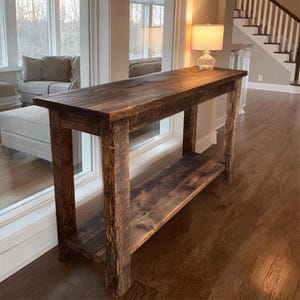 Op de afbeelding: Een rustieke, rechthoekige houten console met een onderste plank. De tafel is gemaakt van donkerbruin, gerecycled hout, met zichtbare nerven en knopen. De tafel is ongeveer 152 cm lang, 38 cm breed en 76 cm hoog.