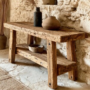 Op de afbeelding: Een rustieke houten bank met een onderste plank, gemaakt van gerecycled hout. De bank heeft een natuurlijke, verweerde afwerking en wordt ondersteund door stevige, vierkante poten. Decoratieve items, waaronder een donkere vaas en een ronde, getextureerde pot, zijn uitgestald.