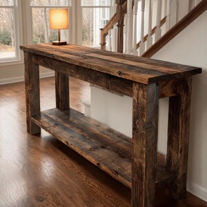 Op de afbeelding: Een rustieke houten console met een onderste plank. De tafel is gemaakt van gerecycled hout met een donkerbruine afwerking. Een kleine lamp met een beige kap staat op het tafelblad. De tafel is ongeveer 152 cm lang.