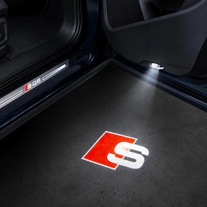 May include: An Audi SQ5 vehicle door sill plate with illuminated branding and a door-mounted projector light casting a red and white Audi S logo onto the ground. The interior door panel features a bright white light beam directed downwards.