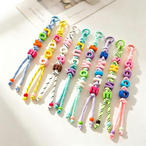 Könnte beinhalten: Acht bunte Schlüsselanhänger aus Kordel mit Kunststoff-Buchstabenperlen, die die Wörter LOVE, GOOD, HOPE, LUCK, MOM, SMILE, SHINE und GOOD bilden. Jeder Anhänger besteht aus gemusterter oder einfarbiger Kordel mit kleinen Akzentperlen an den Enden.