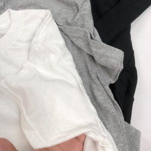 May include: A close-up shot of three folded t-shirts in white, gray, and black. The white t-shirt is in the foreground, with the gray and black shirts layered behind it. The fabric appears soft and lightweight.