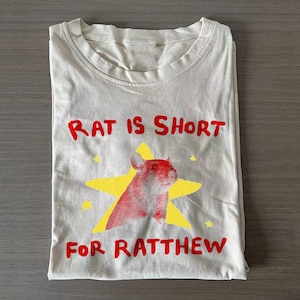 May include: A light beige t-shirt featuring a graphic of a rat centered on a yellow star. The text "RAT IS SHORT" is printed in red above the star, and the text "FOR RATTHEW" is printed in red below the star. The shirt has a crew neckline.