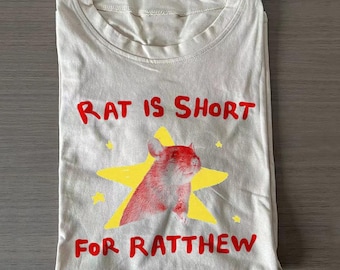 May include: A light beige t-shirt featuring a graphic of a rat centered on a yellow star. The text "RAT IS SHORT" is printed in red above the star, and the text "FOR RATTHEW" is printed in red below the star. The shirt has a crew neckline.
