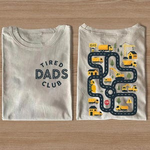 May include: Two folded beige t-shirts. One shirt has the text "TIRED DADS CLUB" in navy blue. The other shirt features a road design with yellow trucks, houses, and trees. The road has a stop sign and a gas station.
