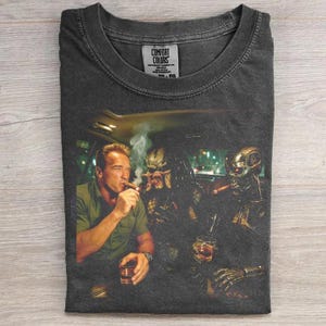 May include: Dark gray Comfort Colors t-shirt featuring a graphic of Arnold Schwarzenegger, Predator, and a Terminator smoking cigars and drinking. The image is set inside a car with city lights in the background.