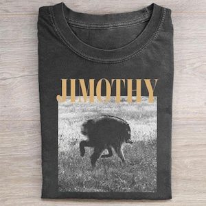 May include: A charcoal gray crew neck t-shirt featuring the word JIMOTHY in large, bold, light tan capital letters above a black and white photographic print of a dog running through a grassy field, centered on the front of the garment.