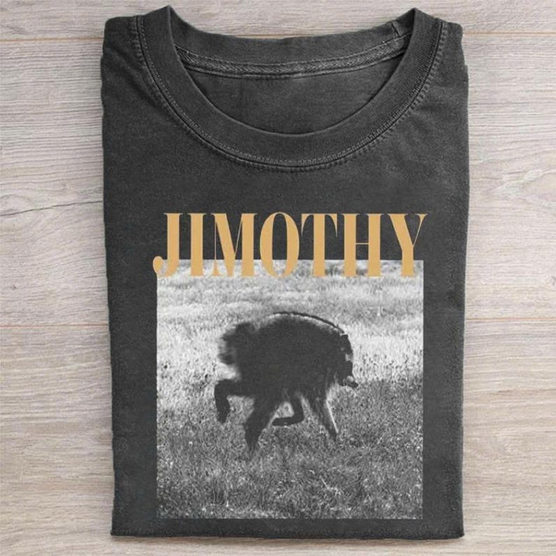 May include: A charcoal gray crew neck t-shirt featuring the word JIMOTHY in large, bold, light tan capital letters above a black and white photographic print of a dog running through a grassy field, centered on the front of the garment.
