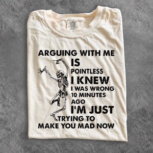 May include: A cream-colored short-sleeve t-shirt featuring a black graphic of a dancing skeleton and the text: ARGUING WITH ME IS POINTLESS I KNEW I WAS WRONG 10 MINUTES AGO I'M JUST TRYING TO MAKE YOU MAD NOW. The shirt has a crew neckline.