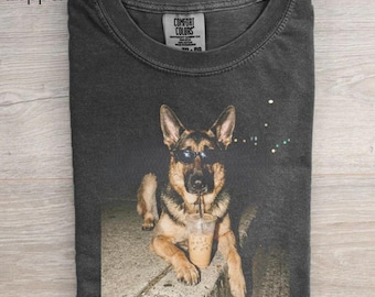 May include: A charcoal gray crew neck t-shirt featuring a graphic print of a German Shepherd dog wearing black sunglasses and drinking from a clear plastic cup with a straw. The inner neck label displays the text Comfort Colors.