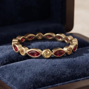 Puede incluir: Una alianza de oro con piedras preciosas rojas de talla marquesa que alternan con piedras amarillas de talla redonda. El anillo se presenta en un estuche de madera forrado de terciopelo azul oscuro con el texto velvetgemstudios en dorado.
